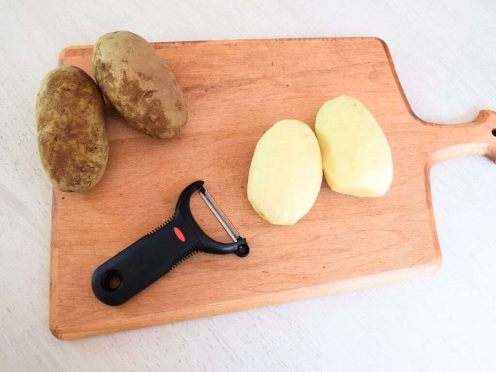 two peeled potatoes and two unpeeled potatoes with a potato peeler on a wooden cutting board