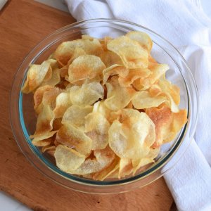 Kettle chips in a glass bowl on a wood cutting board with a white tea towel