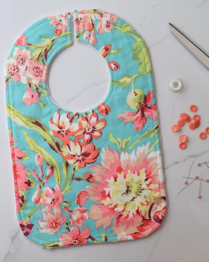 Floral baby bib with snap closure, displayed with sewing tools—thread, scissors, pins, and snaps—on a marbled background.
